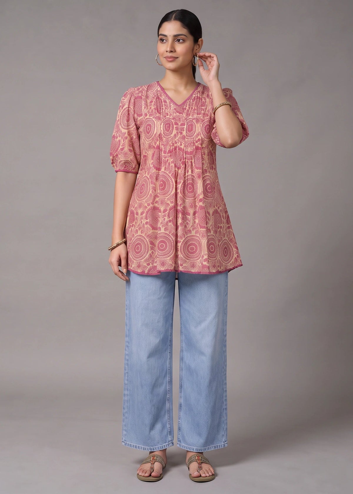 A woman modeling a pink short-sleeve tunic top with a circular floral pattern, paired with wide-leg light blue jeans and brown sandals, against a solid gray studio background.