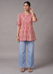 A woman modeling a pink short-sleeve tunic top with a circular floral pattern, paired with wide-leg light blue jeans and brown sandals, against a solid gray studio background.