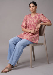 A woman sits on a light beige plastic chair wearing a dusty pink printed kurta with blue jeans and brown sandals, set against a solid gray studio background.