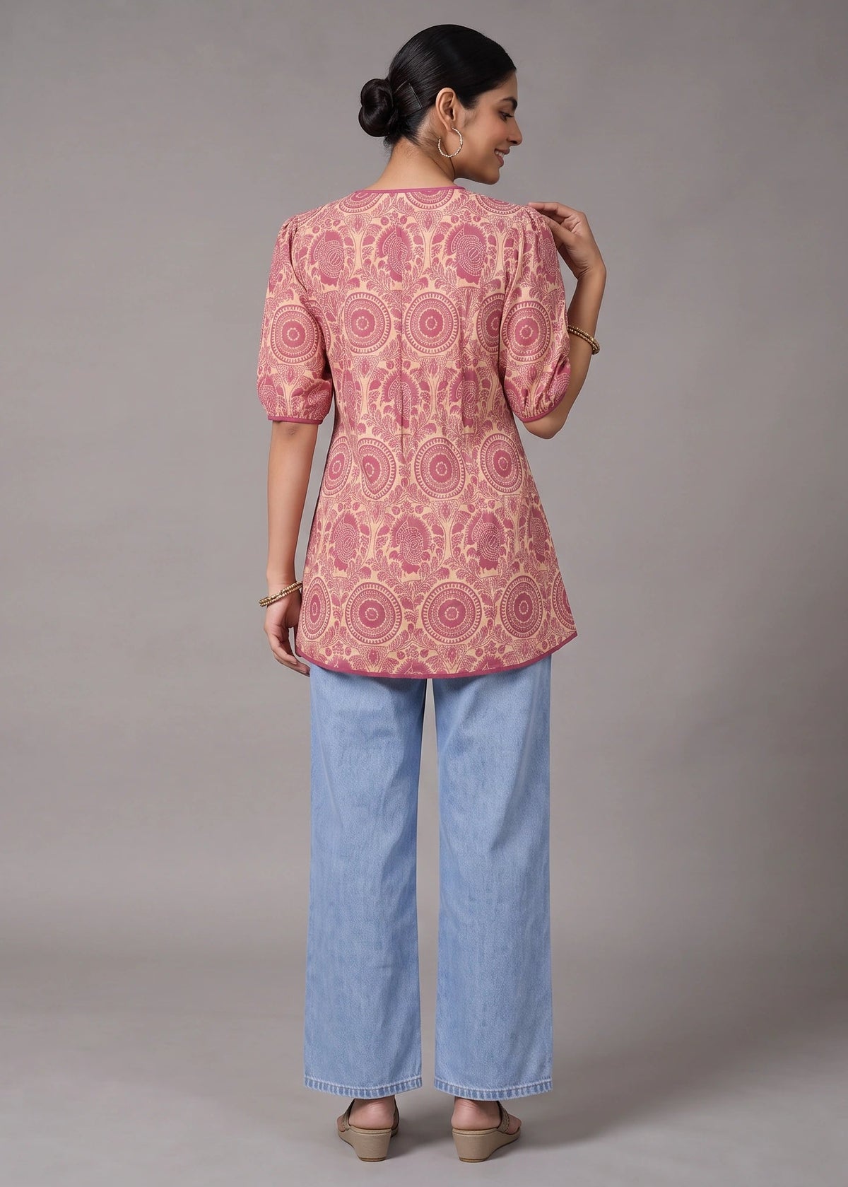 A woman is shown from the back wearing a pink and gold patterned kurta with puff sleeves and light blue wide-leg jeans, standing against a neutral gray studio background.