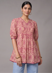 A woman modeling a pink and beige printed tunic top with puff sleeves and a V-neck, paired with light blue jeans, against a solid gray studio background.