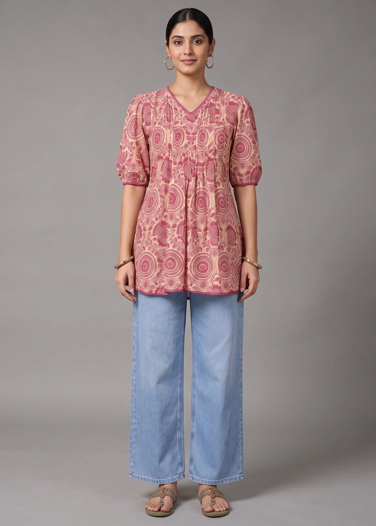 A woman stands against a plain gray studio background wearing a pink patterned short-sleeve tunic top with light blue wide-leg jeans and beige sandals.