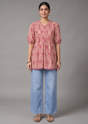 A woman stands against a plain gray studio background wearing a pink patterned short-sleeve tunic top with light blue wide-leg jeans and beige sandals.