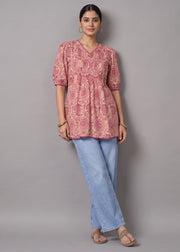 A woman modeling a pink and red patterned short-sleeve tunic top with light blue jeans and beige sandals, standing against a solid gray studio background.