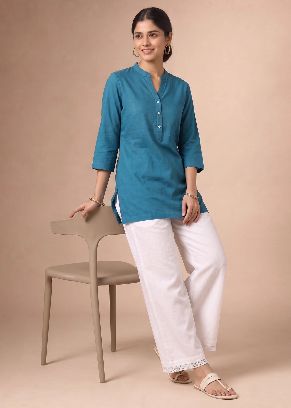 A woman models a teal blue three-quarter sleeve kurta paired with white wide-leg trousers, standing next to a minimalist beige chair against a solid beige studio backdrop.