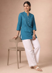 A woman models a teal blue three-quarter sleeve kurta paired with white wide-leg trousers, standing next to a minimalist beige chair against a solid beige studio backdrop.