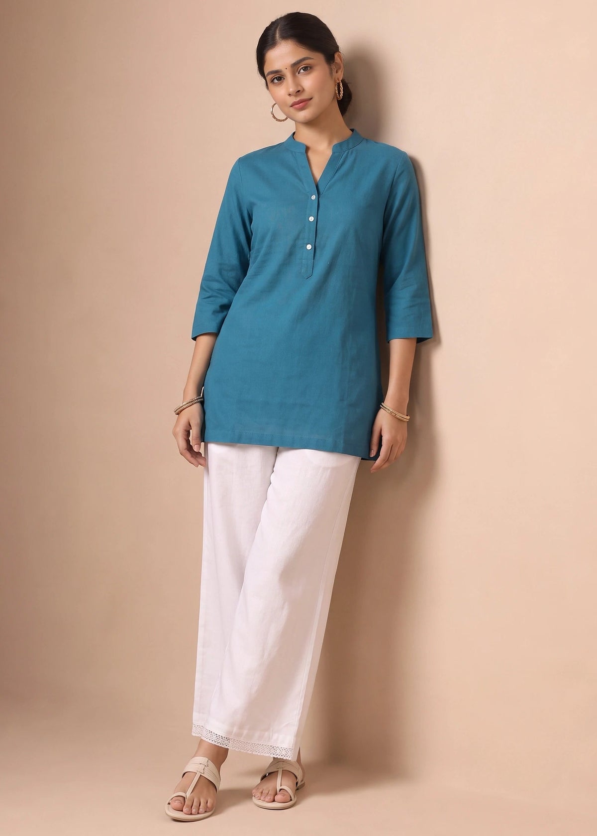 A woman modeling a teal blue three-quarter sleeve kurta with a buttoned mandarin collar, paired with white wide-leg trousers and beige sandals, standing against a solid beige studio backdrop.