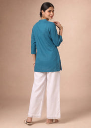A woman wearing a teal blue kurta and white wide-leg trousers, photographed from the back against a solid beige studio backdrop.