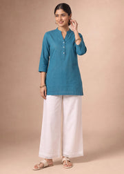 A woman modeling a teal blue three-quarter sleeve kurta with white wide-leg pants and beige sandals, standing against a solid beige studio backdrop.