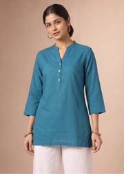 A woman modeling a teal blue linen kurta with a split neckline and three-quarter sleeves, paired with white trousers, against a solid beige studio background.