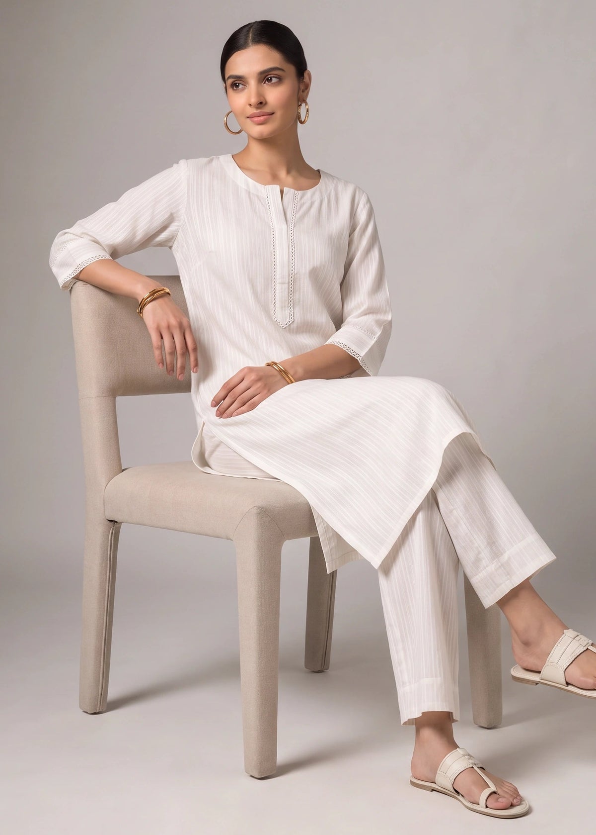 A woman wearing a white linen kurta and matching trousers sits on a beige upholstered chair against a neutral gray studio background.