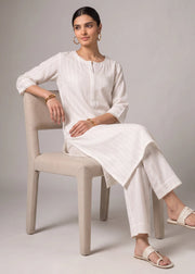 A woman wearing a white linen kurta and matching trousers sits on a beige upholstered chair against a neutral gray studio background.