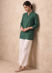 A woman wearing a dark green three-quarter sleeve kurta with a split neckline and white wide-leg trousers, standing against a textured beige plaster wall.
