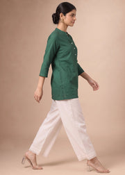 A woman modeling a dark green three-quarter sleeve kurta paired with off-white wide-leg trousers and clear high-heeled sandals, set against a solid beige studio background.