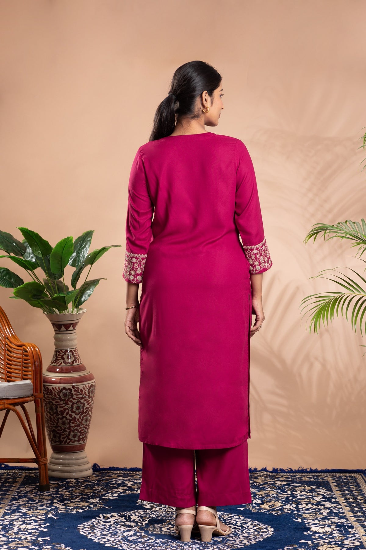 Woman wearing a pink traditional outfit standing against a beige wall with plants and a chair in the background.