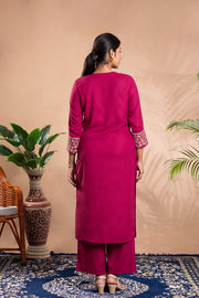 Woman wearing a pink traditional outfit standing against a beige wall with plants and a chair in the background.