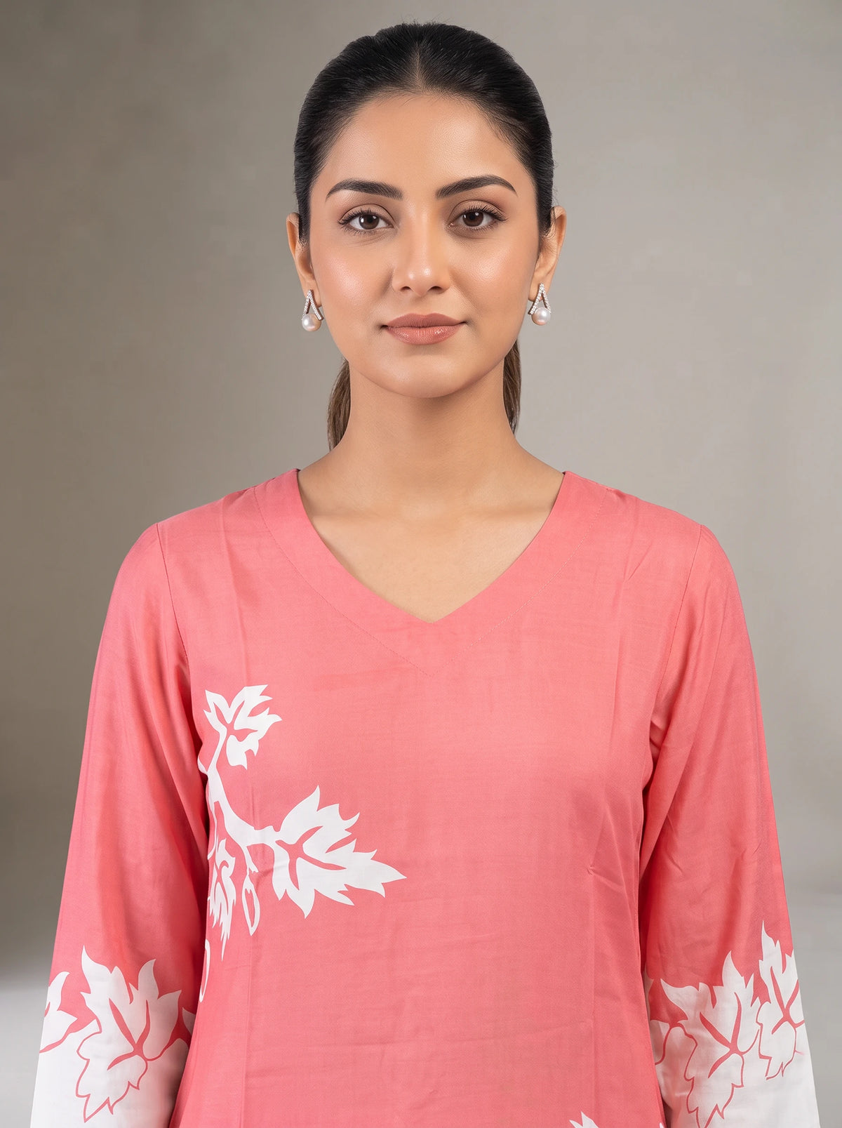 A woman wearing a coral pink V-neck kurta with white leaf embroidery on the chest and cuffs, photographed against a neutral gray studio background.