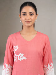 A woman wearing a coral pink V-neck kurta with white leaf embroidery on the chest and cuffs, photographed against a neutral gray studio background.