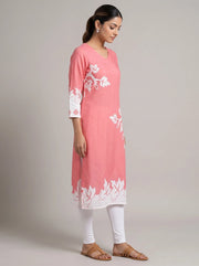 A woman modeling a pink cotton kurta with white floral embroidery, paired with white leggings and brown sandals, set against a plain light gray studio background.