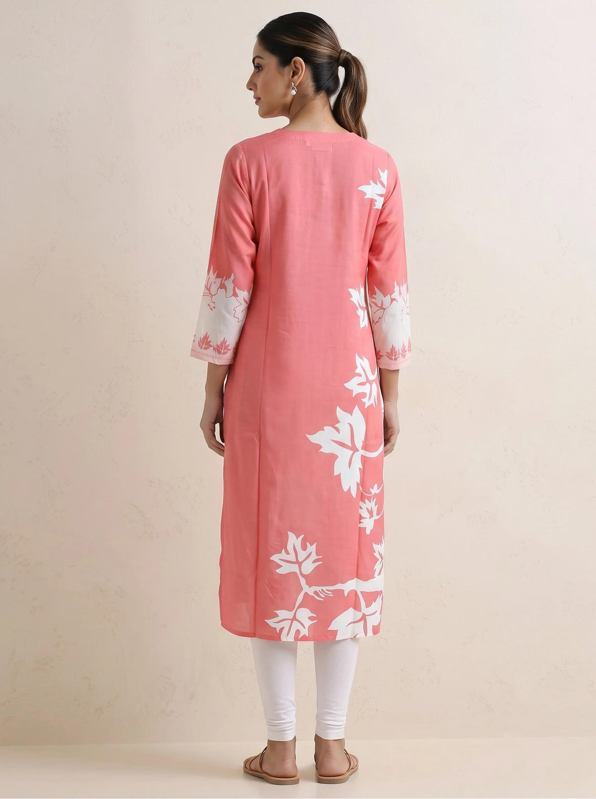 A woman viewed from the back wearing a pink kurta with white floral patterns and white sleeve cuffs, paired with white leggings and brown sandals, against a plain beige studio background.
