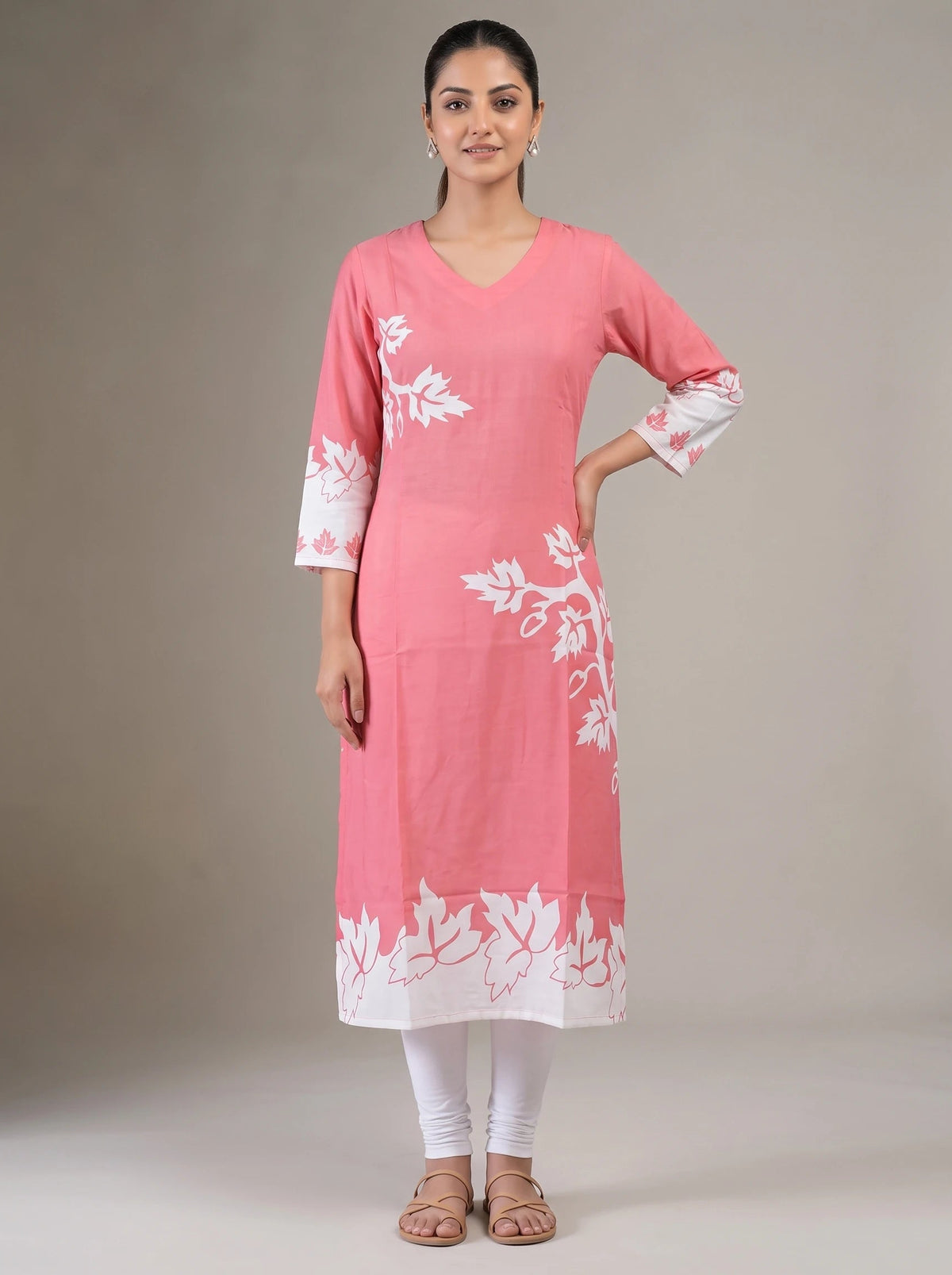 A woman modeling a pink cotton kurta with white floral embroidery and a white border hem, paired with white leggings and brown sandals, against a neutral studio background.
