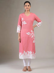 A woman modeling a pink cotton kurta with white floral embroidery and a white border hem, paired with white leggings and brown sandals, against a neutral studio background.