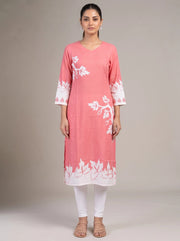 A woman modeling a pink cotton kurta with white floral embroidery and white lace trim on the sleeves and hem, paired with white leggings and beige sandals, against a neutral studio background.