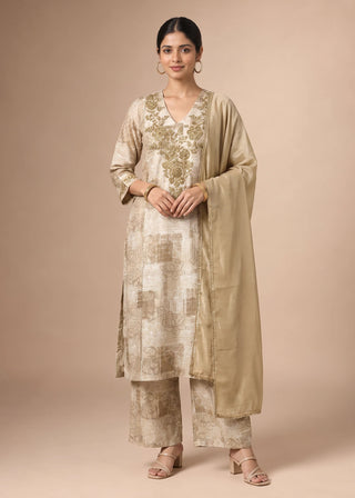 A woman modeling a traditional Indian outfit consisting of a beige kurta with gold floral embroidery, matching trousers, and a sheer beige dupatta, set against a solid beige studio background.