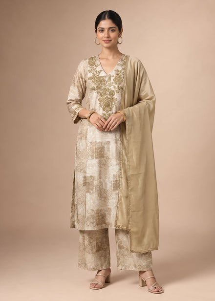 A woman modeling a traditional Indian outfit consisting of a beige kurta with gold floral embroidery, matching trousers, and a sheer beige dupatta, set against a solid beige studio background.