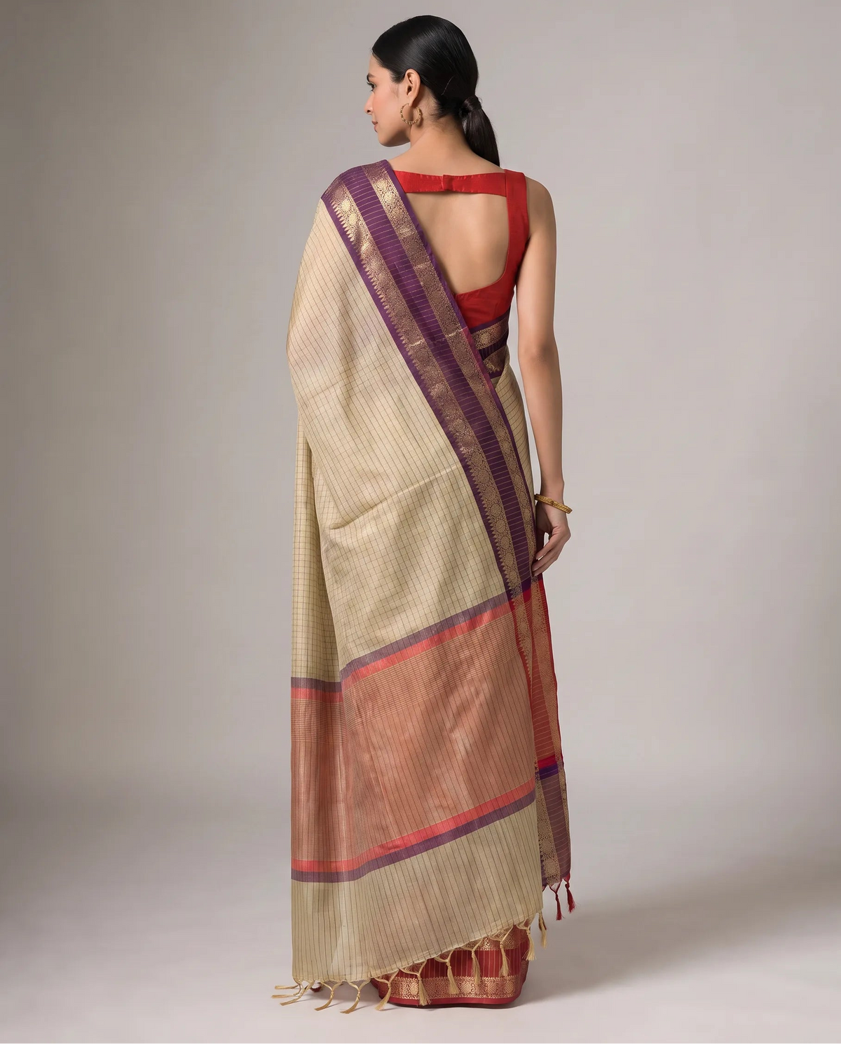A woman shown from the back wearing a beige and rust-orange striped saree with a purple border and a red blouse, photographed against a neutral gray studio background.