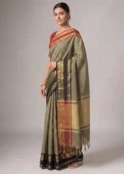 A woman modeling a traditional Indian saree with a muted olive green body, black and gold stripes, and a red and gold border, set against a plain gray studio background.