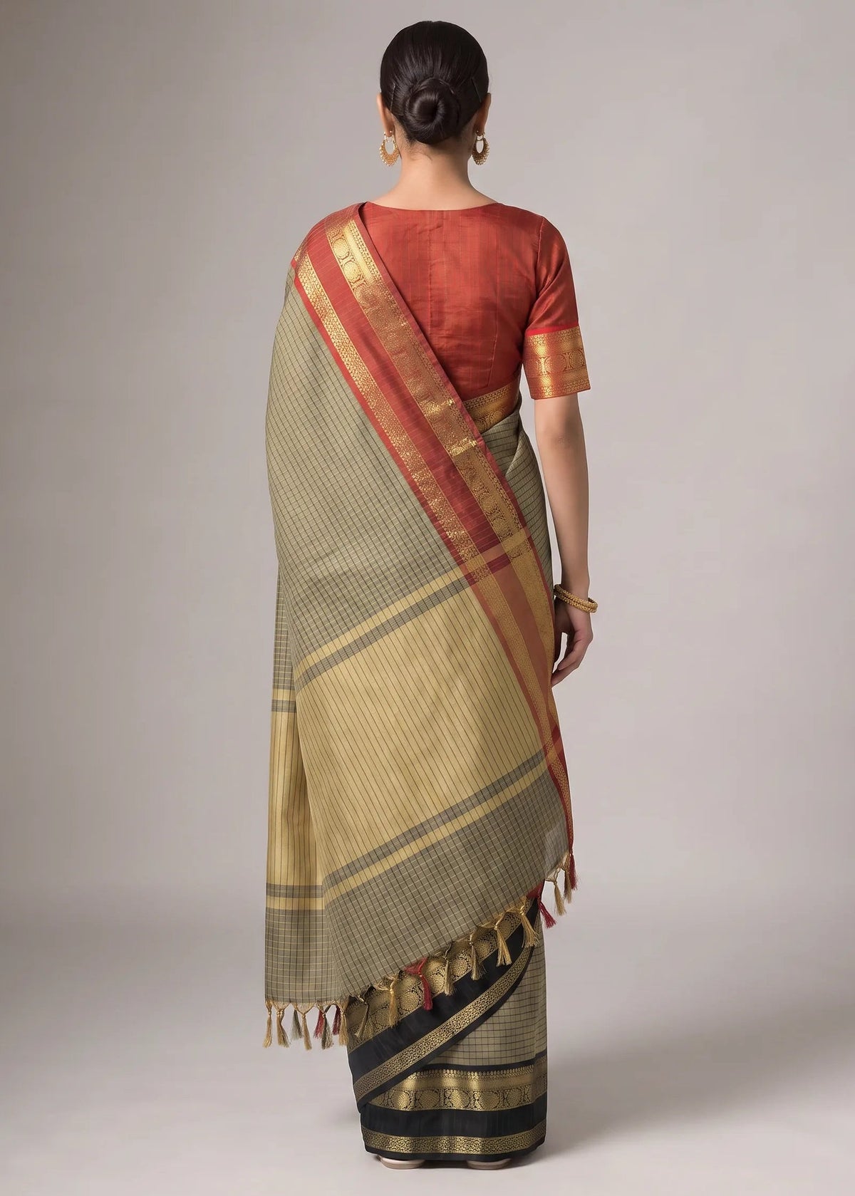 A woman shown from the back wearing a traditional Indian saree. The saree has a beige body with a red border and gold zari detailing, paired with a rust-red short-sleeve blouse.