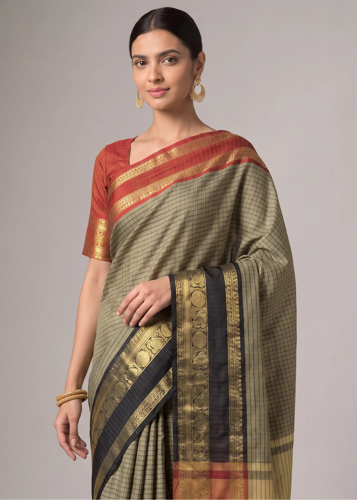 A woman modeling a traditional Indian saree with a muted olive green body, a red and gold patterned blouse, and a black and gold border, set against a plain gray studio background.