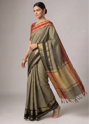 A woman models a traditional Indian saree with a textured olive green body, black and gold borders, and a red and gold pallu, set against a plain light gray studio background.