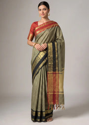 A woman modeling a traditional Indian saree with a muted olive green body, black and red borders, and gold zari detailing, paired with a red blouse.