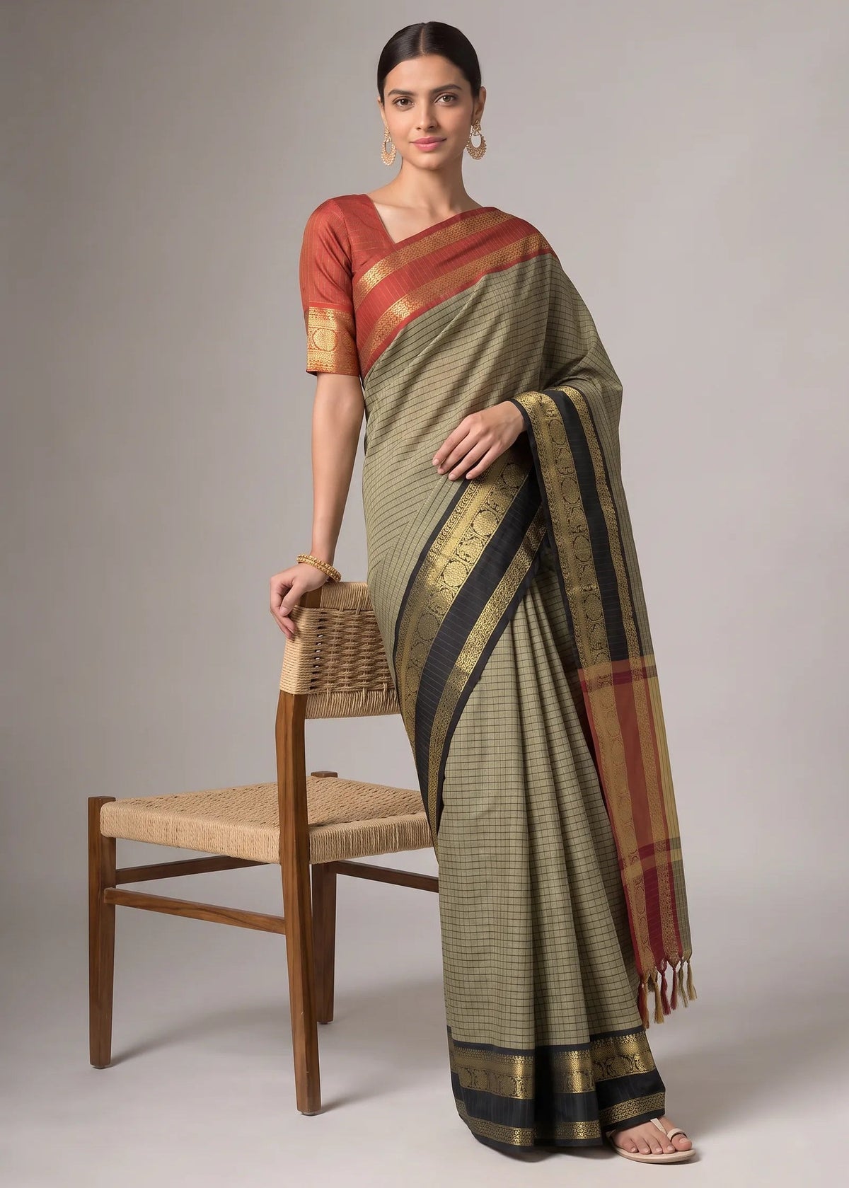 A woman models a traditional Indian saree in a studio setting, standing next to a wooden chair. The saree has a muted olive green body with a fine black checkered pattern, a rust-red and gold border on the pallu, and a dark navy and gold zari border.