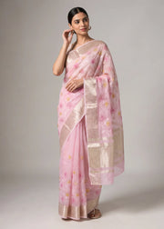 A woman modeling a light pink saree with a metallic beige border and scattered floral motifs, standing against a neutral gray studio background.