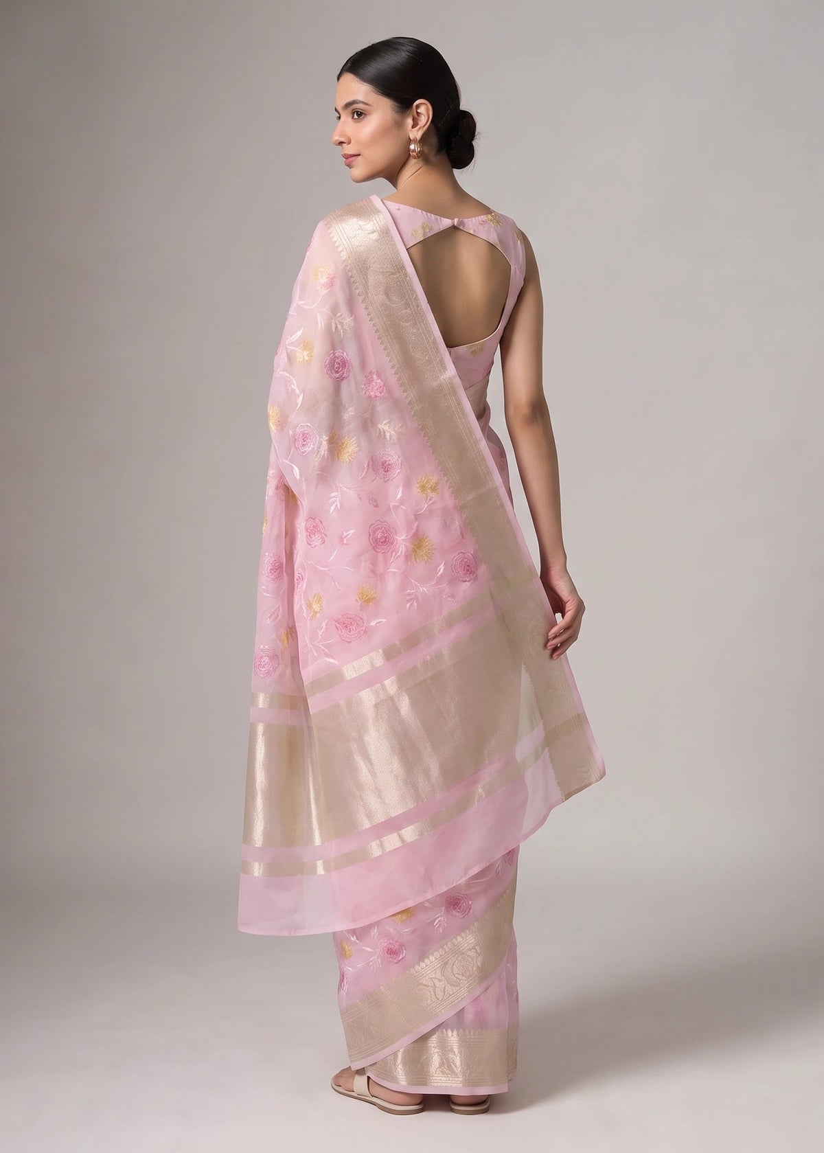 A woman modeling a light pink and beige floral saree with metallic gold borders, shown from the back against a neutral gray studio background.