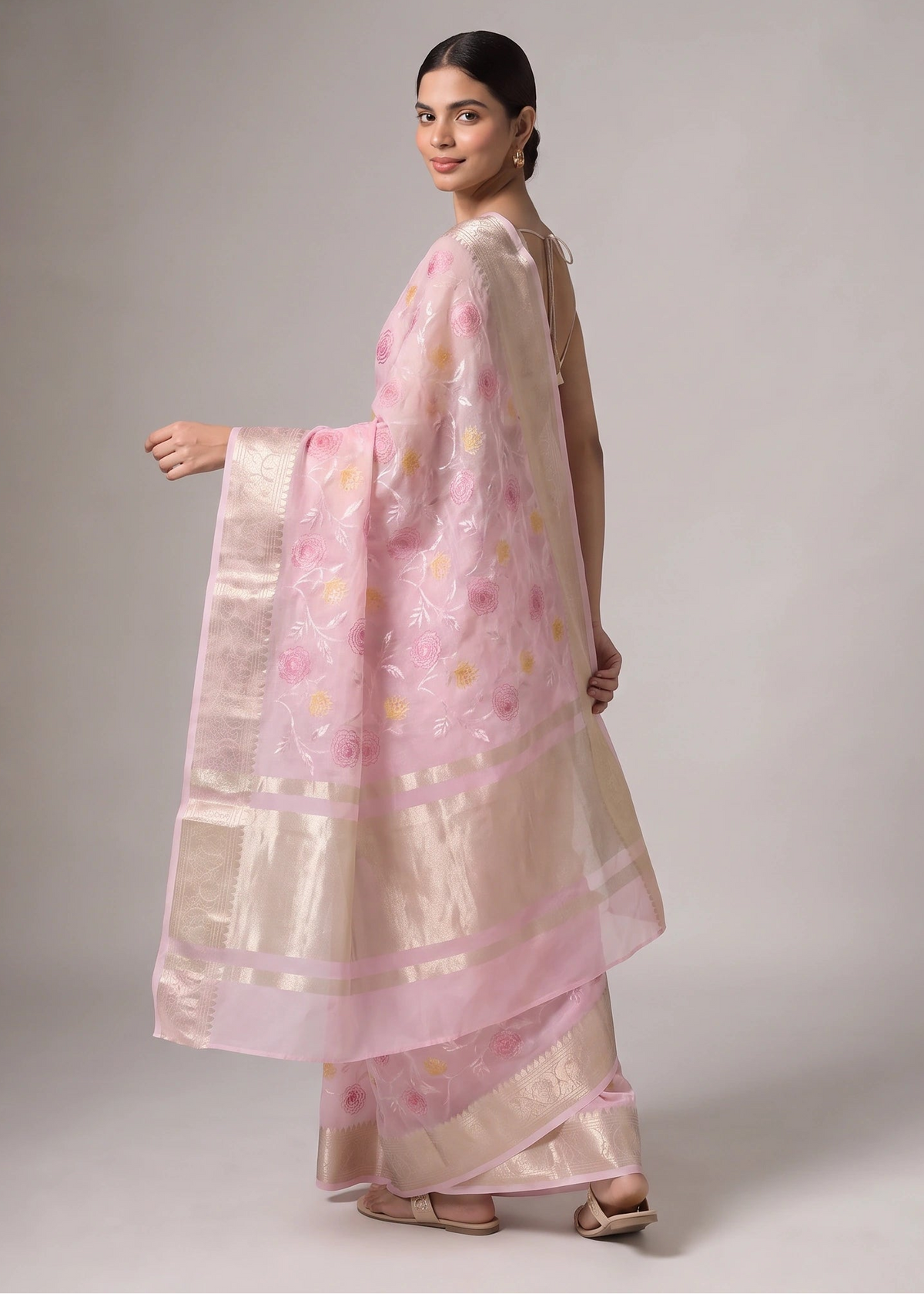 A woman modeling a light pink and beige saree with floral embroidery and a wide metallic border, photographed against a neutral gray studio background.