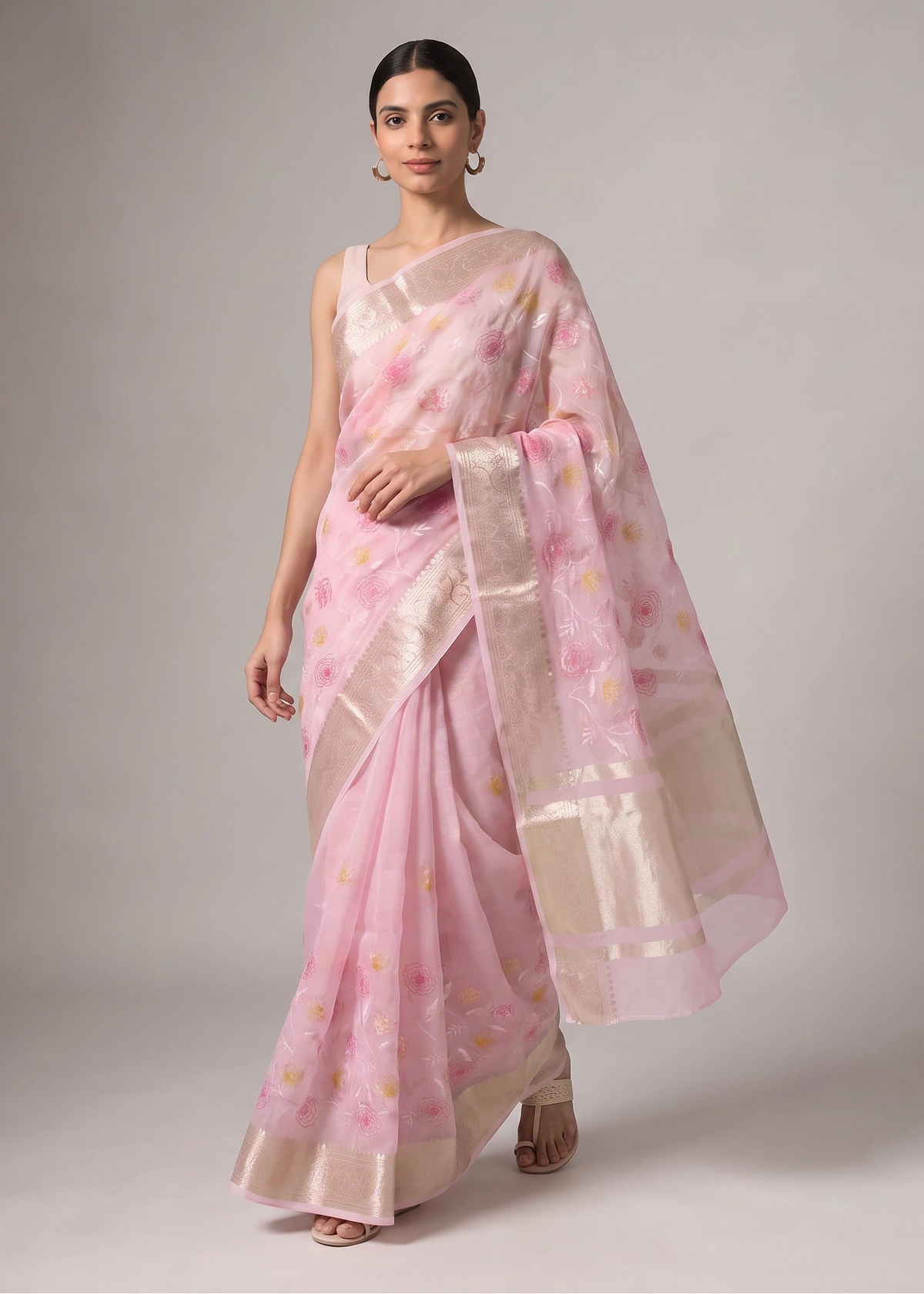 A woman models a light pink saree with a subtle floral pattern and a wide beige border, set against a neutral gray studio background.