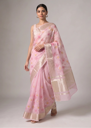 A woman models a light pink saree with a subtle floral pattern and a wide beige border, set against a neutral gray studio background.