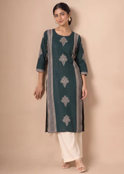 A woman modeling a dark teal green kurta with silver-gray floral embroidery and striped details, paired with white pants, against a solid beige studio background.