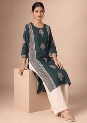 A woman models a dark teal-green kurta with gray and white embroidered patterns, paired with white pants, sitting on a beige block against a solid beige background.