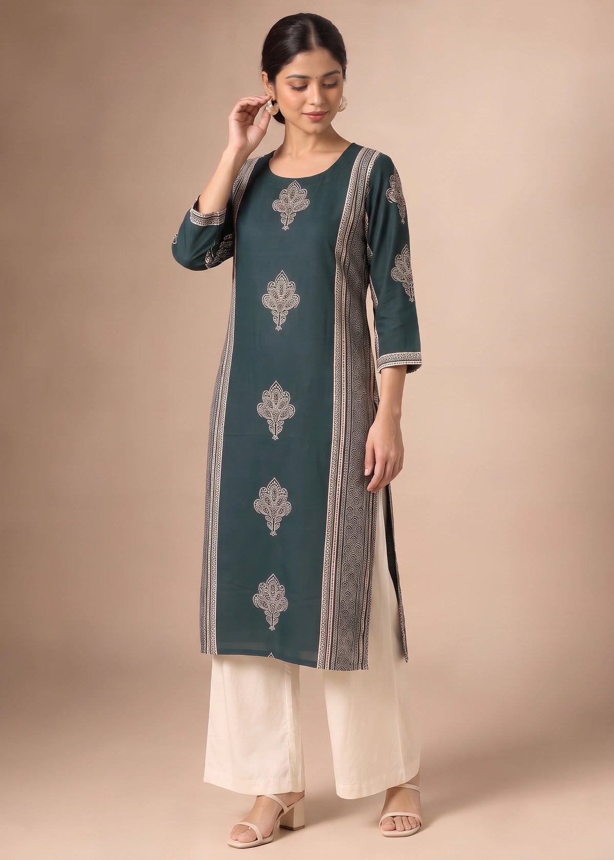 A woman modeling a dark teal green kurta with silver-gray floral embroidery and striped borders, paired with off-white wide-leg pants, against a solid beige studio background.