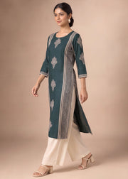 A woman modeling a dark teal green kurta with silver-gray printed motifs and striped borders, paired with cream-colored palazzo pants, against a solid beige studio background.