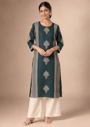 A woman modeling a dark teal green kurta with silver-gray printed motifs and vertical stripes, paired with off-white wide-leg pants, against a solid beige studio background.