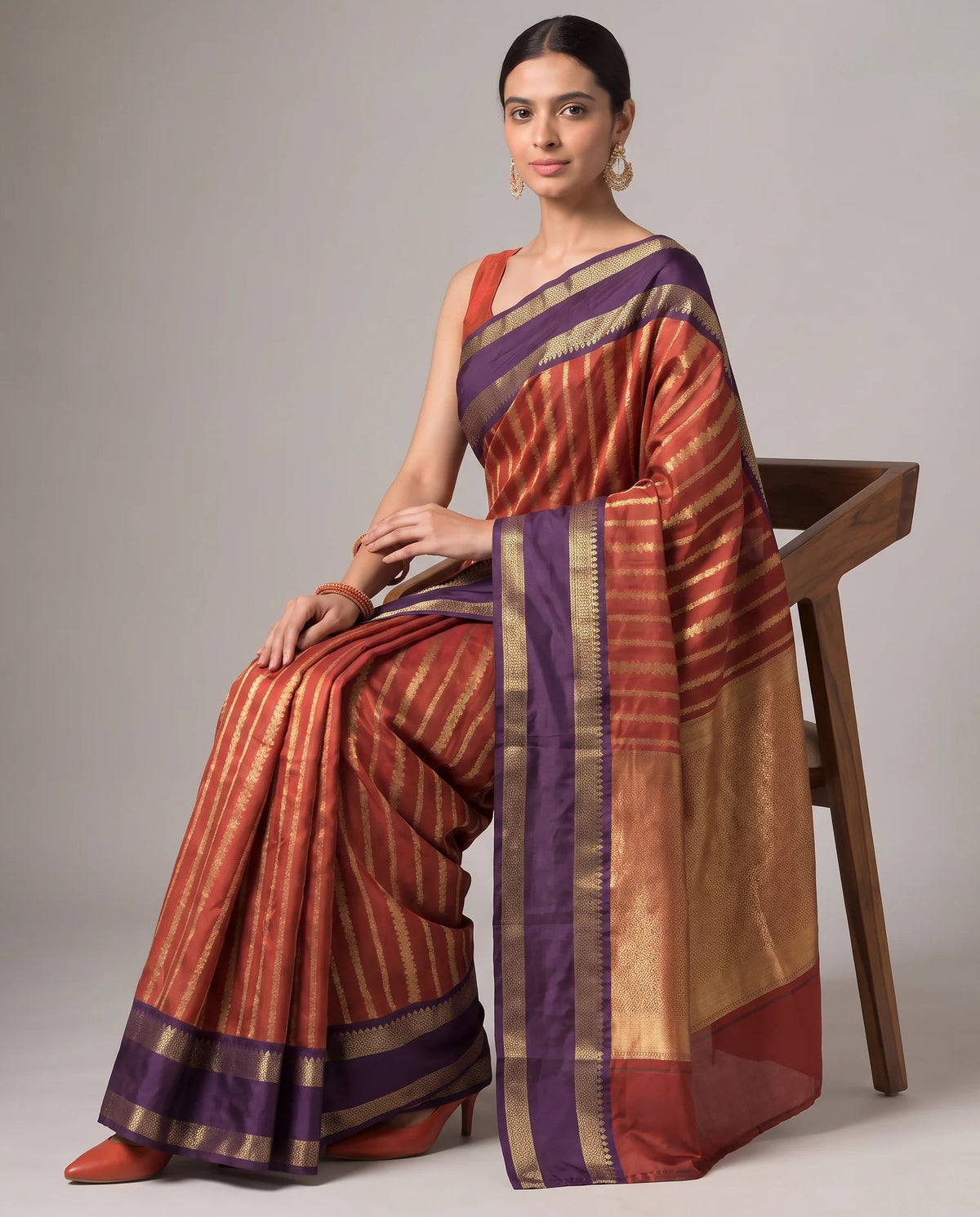 A woman seated on a wooden chair wearing a draped orange and gold striped silk saree with purple borders, photographed against a plain light gray studio background.