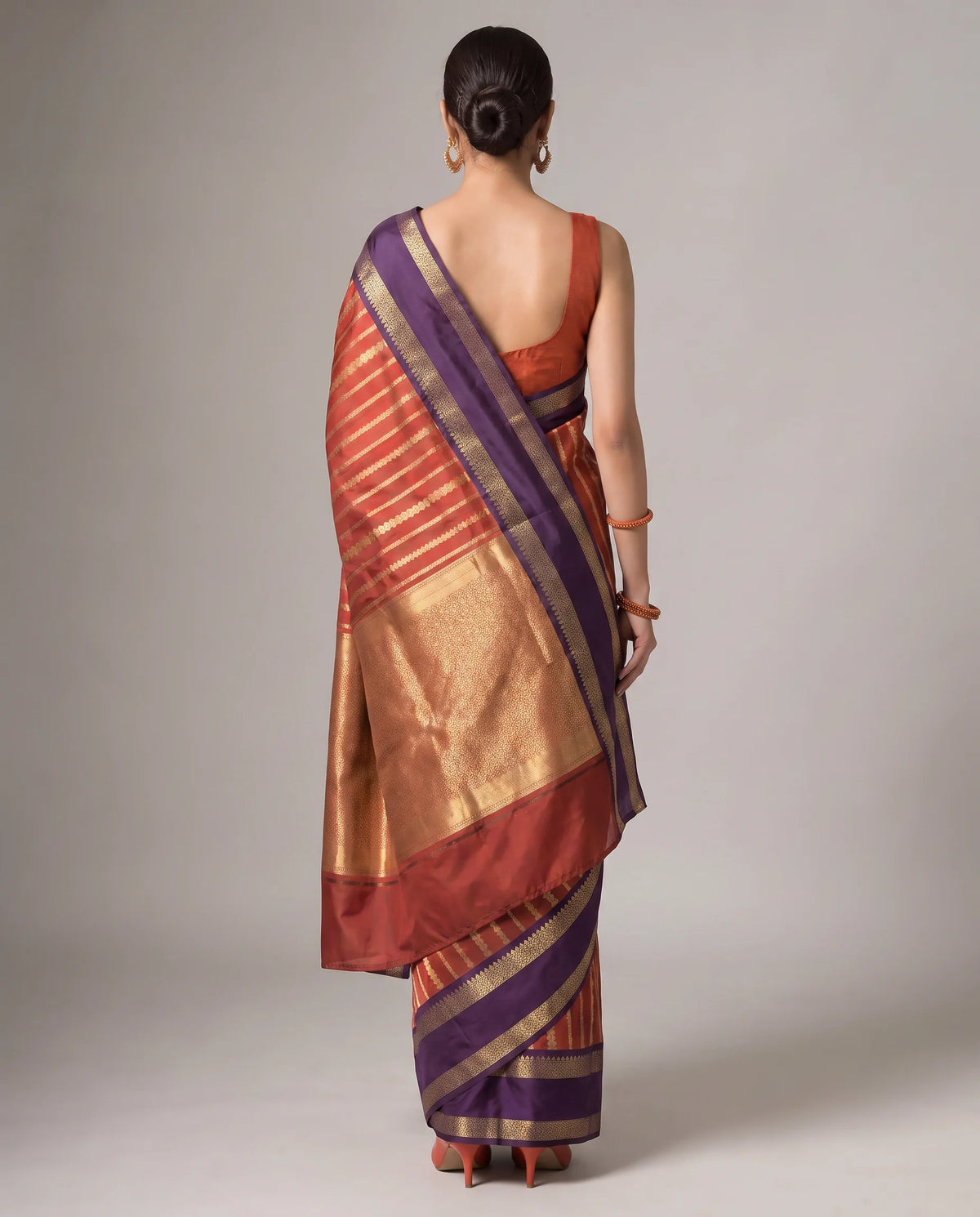 A woman shown from the back wearing a traditional Indian saree. The saree features a rich color palette of deep purple, metallic gold, and terracotta red with fine horizontal stripes.