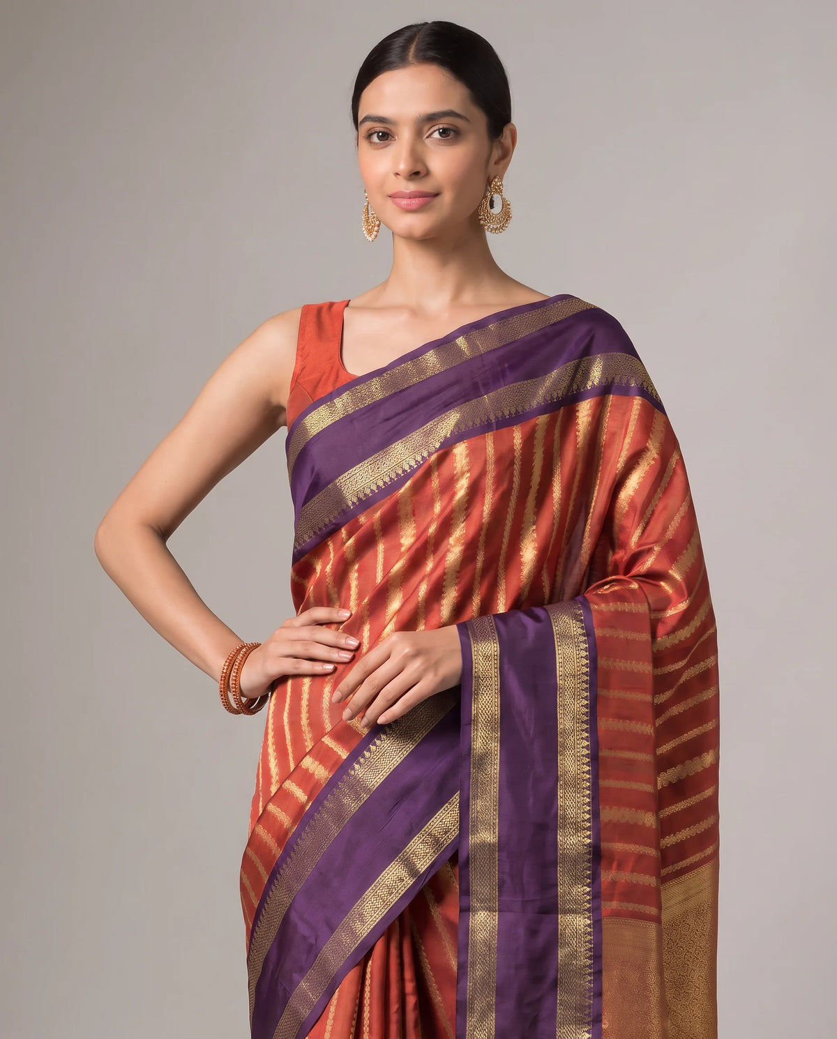 A woman modeling a purple and rust-orange striped silk saree with gold zari borders, paired with a matching rust-orange blouse, against a solid gray studio background.