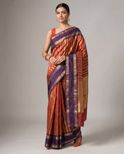 A woman modeling a traditional Indian silk saree with a copper-orange body, purple borders, and gold zari metallic stripes, set against a plain gray studio background.